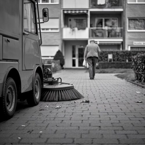 Kehrmaschine reinigt die Straße in einer Wohnanlage mit Betonfassaden; ein Bewohner geht vorbei.